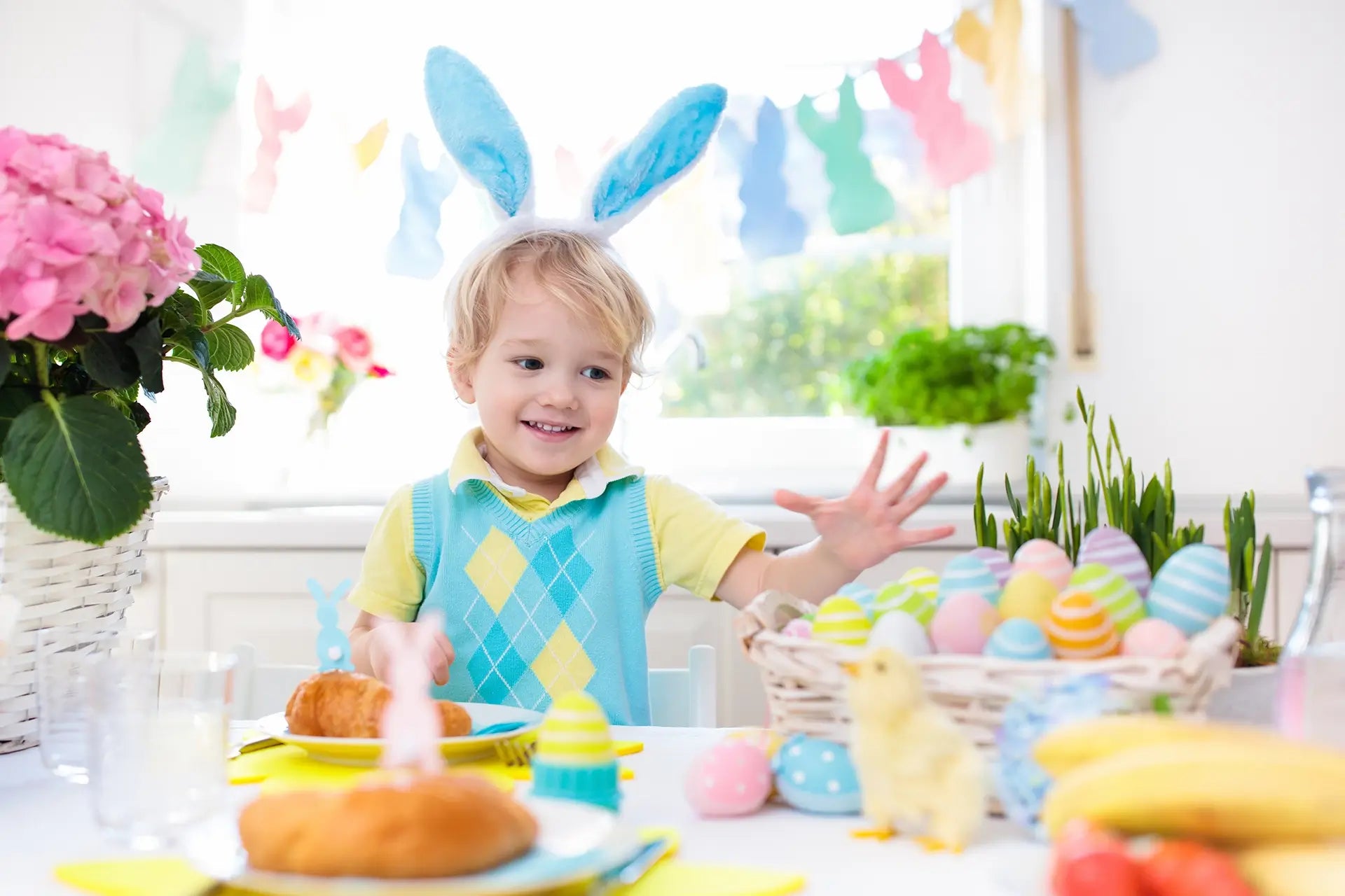 A cheerful young boy wearing blue bunny ears enjoys an Easter celebration, surrounded by colorful eggs, a chick, and festive decorations.