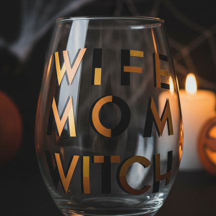 Close up of Celebrate It Wife Mom Witch stemless wine glass showing bold black and gold lettering detail with moody candlelit Halloween background