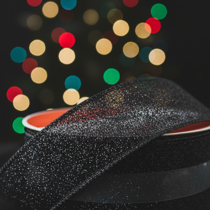 Close up of solid black glitter wired ribbon showing glitter texture and sheen detail with dark moody bokeh Christmas lights background