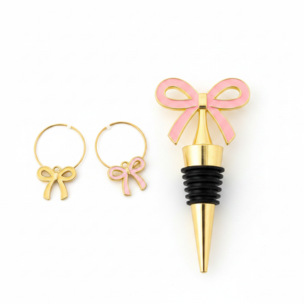 Close-up of gold bottle stopper with pink bow charm and two gold wine glass charms with bow designs on white background