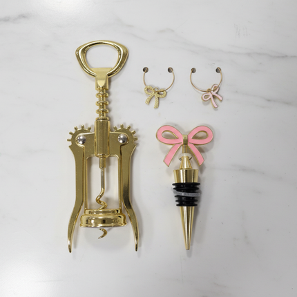 Gold corkscrew, gold bottle stopper with pink bow charm, and two gold wine glass charms laid out on a white marble surface
