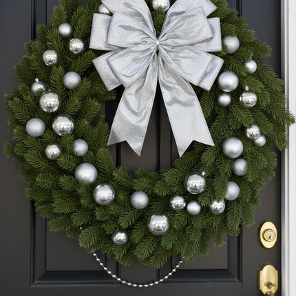 Large silver glitter decorative bow attached to a Christmas wreath with pine branches and silver ornaments hanging on a front door