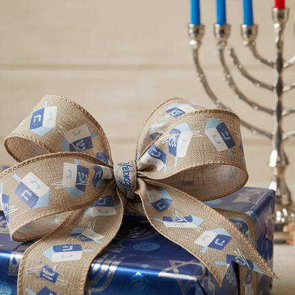 Celebrate It Hanukkah wired ribbon with dreidel pattern tied as a bow on a gift wrapped in blue and silver paper with menorah in background